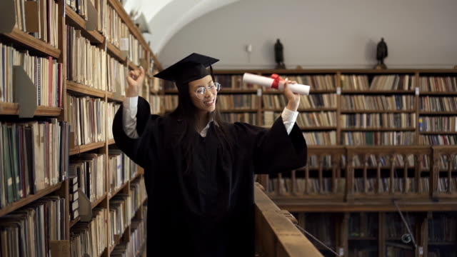 Attractive-student-in-academic-dress-is-having-fun,-standing-in-library