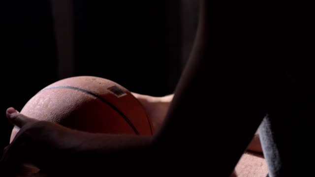 Jugador-de-baloncesto-sentado-y-sosteniendo-la-bola,-esperando-al-juego-empezar,-el-fondo-de-la-habitación-oscura