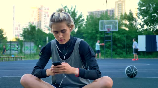 Beautiful-young-woman-sitting-on-basketball-court-and-typing-on-phone,-wearing-earphones,-park-with-skyscrapers-in-background