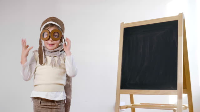 Ein-kleines-Mädchen-gekleidet-wie-ein-Pilot-oder-Pilotin,-zeigt-mit-der-Hand-die-Tafel-hinter-ihr-wie-ein-Flug-Insign-zu-lernen,-Flugzeuge-und-Phantasie-zu-nutzen.
