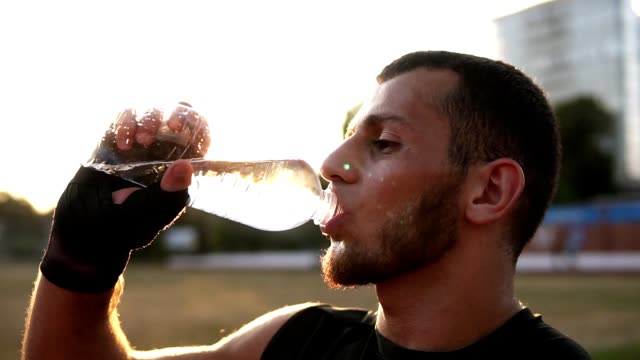Side-view-of-a-young-male-boxer-in-black-bandages-has-a-rest-during-the-training-outdoors,-drinking-water-from-a-plastic-bottle.-Sun-shines-on-the-background