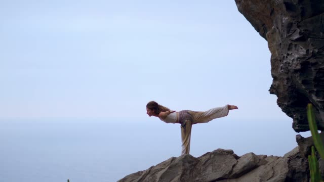 Woman-meditating-in-yoga-warrior-pose-at-the-ocean,-beach-and-rock-mountains.-Motivation-and-inspirational-fit-and-exercising.-Healthy-lifestyle-outdoors-in-nature,-fitness-concept.