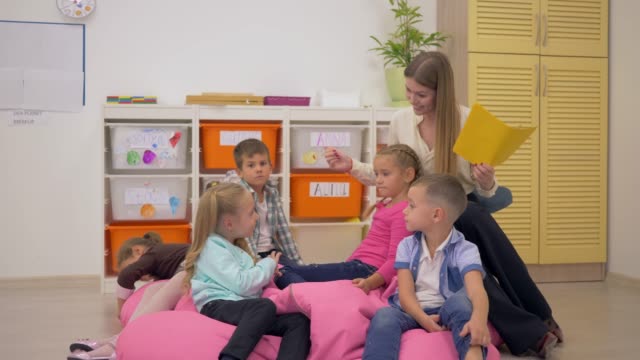 freundliche-Lehrerin-auf-Bohnenbeutelstuhl-sitzen-und-sprechen-mit-kleinen-Kindern-im-Klassenzimmer-in-der-Schule