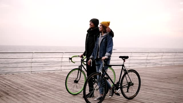 Hermosa-pareja-sonriente-de-los-hipsters-jóvenes-caminando-juntos-abrazando-con-sus-bicicletas-cerca-del-mar-en-día-de-otoño.-Chica-joven-de-sombrero-amarillo-en-algo.-Caminando-por-cubierta-de-madera-durante-el-día.-Horizonte-de-la-mar