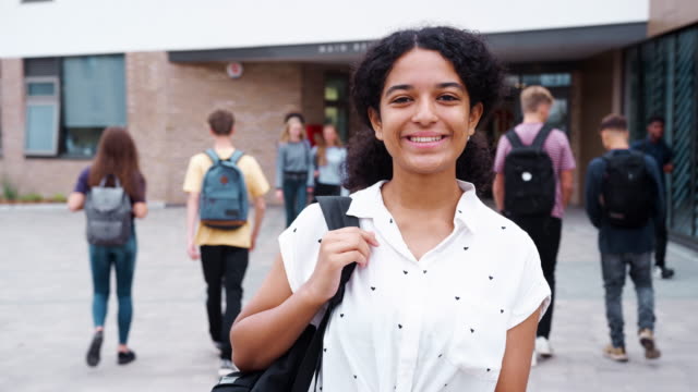 Portrait-Of-Smiling-Female-High-School-Student-Outside-College-Building-With-Other-Teenage-Students-In-Background