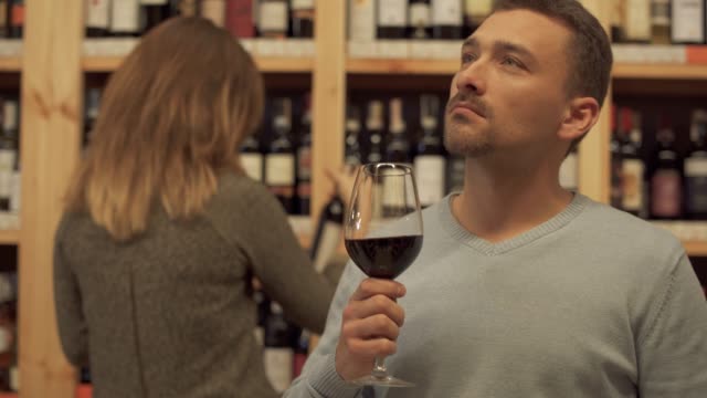 Handsome-pensive-guy-with-a-wine-glass-in-a-liquor-store.-Woman-in-the-background-is-choosing-a-wine-bottle.