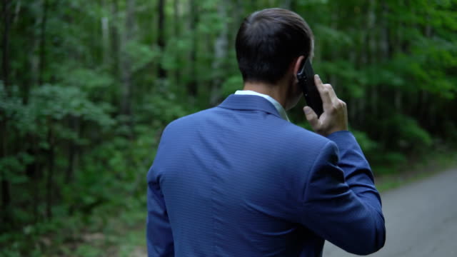 handsome-man-walking-along-the-road-in-forest-talking-on-the-phone,-view-back