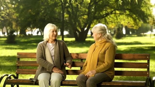 Two-grandmothers-talking,-friendly-sitting-on-bench-in-park,-old-golden-years