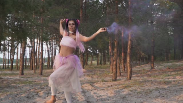 Beautiful-lady-with-bright-makeup-in-a-pink-dress-dancing-with-smoke-bombs-on-the-background-of-pine-trees.-The-dance-of-a-sensual-woman-with-a-flower-hairstyle.-Slow-motion.