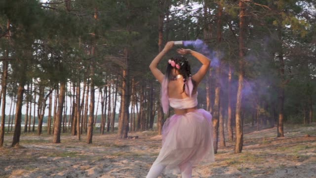 Charmante-Frau-mit-hellen-Make-up-in-einem-rosa-Kleid-tanzen-mit-Rauchbomben-auf-dem-Hintergrund-der-Pinien.-Der-Tanz-von-einem-sinnlichen-Mädchen-mit-einer-wunderschönen-Frisur-mit-Blumen.-Slow-Motion.