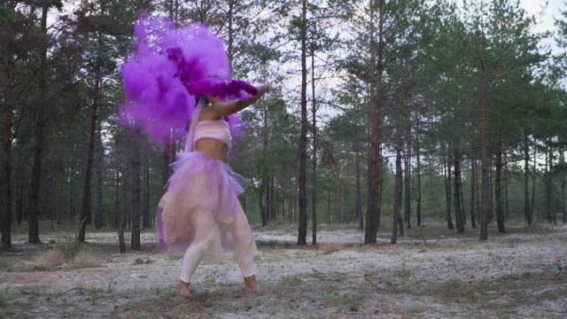 Zärtliche-Frau-mit-hellen-Make-up-in-einem-rosa-Kleid-Tanz-mit-brennenden-Rauchbomben-auf-dem-Hintergrund-der-Pinien.-Der-Tanz-von-einem-sinnlichen-Mädchen-mit-einer-wunderschönen-Frisur-mit-Blumen.-Slow-Motion.