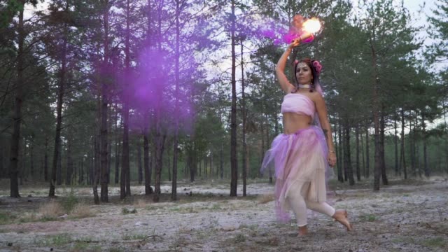 Cute-young-woman-with-bright-makeup-in-a-pink-dress-dancing-with-burning-smoke-bombs-on-the-background-of-pine-trees.-The-dance-of-a-sensual-girl-with-a-flower-hairstyle.-Slow-motion.