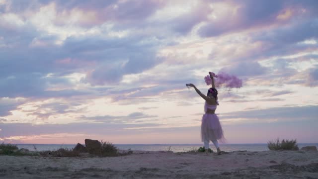 Tender-girl-with-sparkling-makeup-in-a-pink-dress-dancing-with-smoke-bombs-on-the-bank-of-the-river.-The-dance-of-a-sensual-woman-with-a-wonderful-hairstyle-with-flowers.-Slow-motion.
