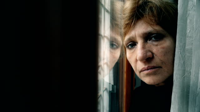 Sad-and-depressed-mature-woman-leaning-against-the-window,-looks-at-the-camera