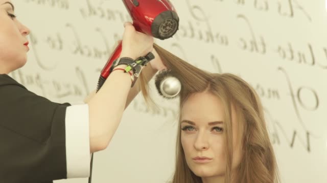 Blonde-Frau-bei-Frisuren-lange-Haare-mit-Trockner-und-Haarbürste-im-Friseursalon.-Nahaufnahme-Haircutter-Frau-Haare-mit-Trockner-trocknen-und-Kämmen-nach-Waschen-und-schneiden