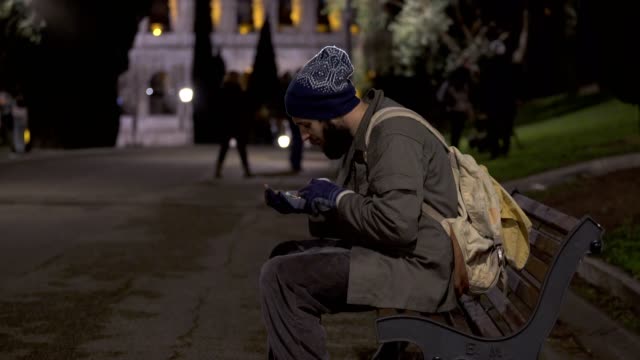 Lonely-beggar-sit-on-bench-at-night,-cheking-his-alms