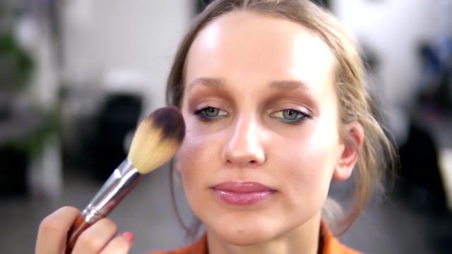 Front-view-of-a-beautiful-smiling-female-model-during-the-make-up-process.-Make-up-artist-completes-the-work,-putting-some-powder-using-a-brush.-Close-up-footage