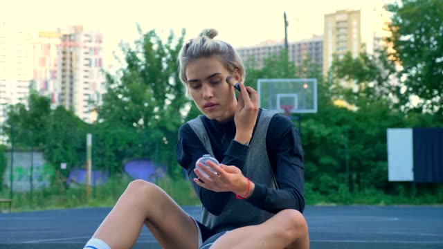Attraktiven-weiblichen-Basketball-Spieler-sitzen-auf-Hof-und-tun-Make-up,-Parks-und-Gebäude-im-Hintergrund
