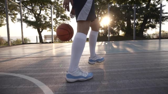 Close-up-footage-of-a-female-athlete-legs-in-white-golf-socks-and-sneakers.-Female-baasketball-player-bouncing-ball-from-hand-to-hand.-Sun-shines-on-the-background