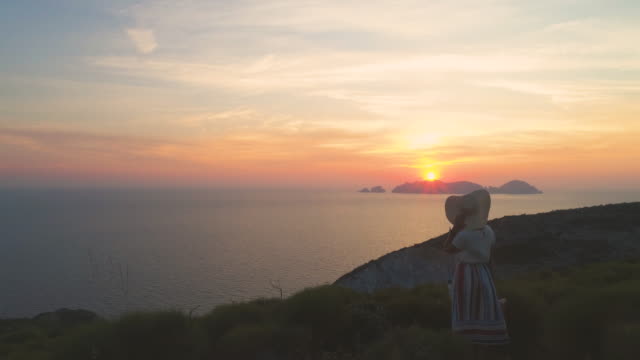 Hermosa-mujer-joven-con-sombrero-de-moda-colorido-vestido-con-la-falda-y-flores-mirando-la-puesta-de-sol-en-montaña-de-la-isla-de-Ponza-Italia.-Tiro-de-Drone-aéreo.