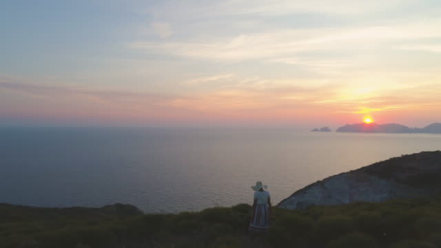 Schöne-junge-Frau-mit-Hut-in-der-Mode-bunt-Kleid-mit-Rock-und-Blick-auf-den-Sonnenuntergang-auf-Ponza-Inselberg-Italien-Blumen.-Luftaufnahme-der-Drohne.