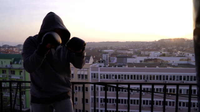 Fighter-with-hood-on-head-boxing-on-terrace-in-sunset.-man-punching-boxing-bag,-sportsman-practicing,-power-training,-strong-guy-hard-exercising,-strength-exercises,-workout,-handheld,-sunny-day.