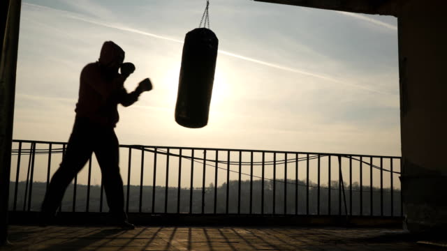 Silhouette-of-man-boxer-while-punches-boxing-bag.-Beautiful-sunset-in-background,-sportsman-practicing,-power-training,-strong-guy-hard-exercising,-strength-exercises,-workout,-handheld,-sunny-day.