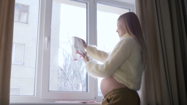 Waiting-of-baby,-young-pregnant-female-with-big-tummy-looks-at-little-children-clothes-for-future-baby-against-window-in-sunlight
