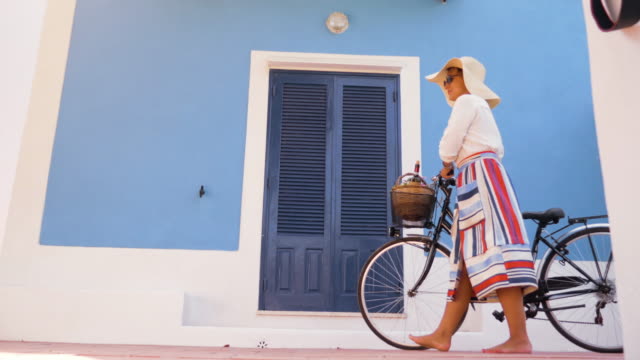 Mujer-joven-feliz-con-bicicleta-caminando-frente-a-patio-de-la-puerta-de-la-casa-azul.-Camisa-de-moda-blanco,-gran-sombrero,-falda-colorida-y-gafas-de-sol.-Isla-de-Ponza,-Italia.