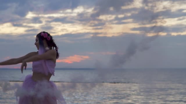 Nettes-Mädchen-mit-hellen-Make-up-in-einem-rosa-Kleid-tanzen-mit-Rauchbomben-am-Ufer-des-Flusses.-Der-Tanz-einer-sinnlichen-Frau-mit-einer-wunderschönen-Frisur-mit-Blumen.-Slow-Motion.