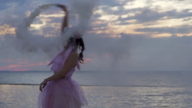 Tender-girl-with-sparkling-makeup-in-a-pink-dress-dancing-with-smoke-bombs-on-the-bank-of-the-river.-The-dance-of-a-sensual-woman-with-a-wonderful-hairstyle-with-flowers.-Slow-motion.