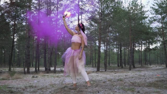 Cute-tender-woman-with-bright-makeup-in-a-pink-dress-dancing-with-burning-smoke-bombs-on-the-background-of-pine-trees.-The-dance-of-a-sensual-girl-with-a-flower-hairstyle.-Slow-motion.