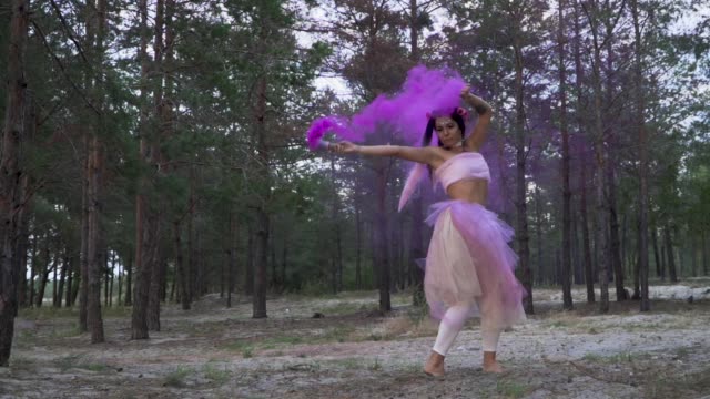 Tender-woman-with-bright-makeup-in-a-pink-dress-dancing-with-burning-smoke-bombs-on-the-background-of-pine-trees.-The-dance-of-a-sensual-girl-with-a-wonderful-hairstyle-with-flowers.-Slow-motion.