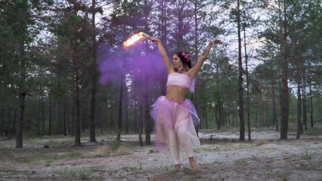 Cute-tender-woman-with-bright-makeup-in-a-pink-dress-dancing-with-burning-smoke-bombs-on-the-background-of-pine-trees.-The-dance-of-a-sensual-girl-with-a-flower-hairstyle.-Slow-motion.