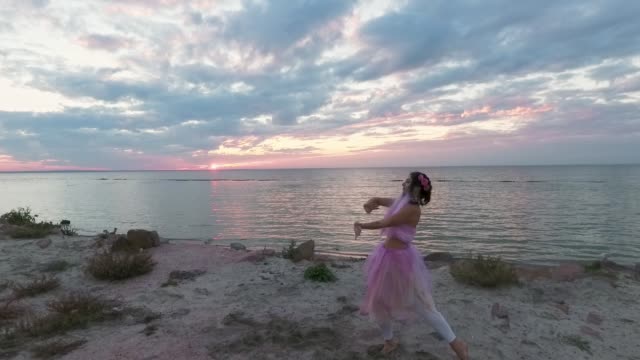 Atop-view-of-sensual-young-woman-with-sparkling-makeup-in-a-pink-dress-dancing-with-smoke-bombs-on-the-bank-of-the-river.-The-dance-of-a-pretty-sweet-girl.-Slow-motion.-Shooting-from-the-drone.