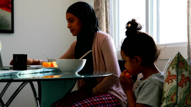 Madre-joven-usando-laptop-mientras-su-hija-está-comiendo-frutas-en-casa-4k