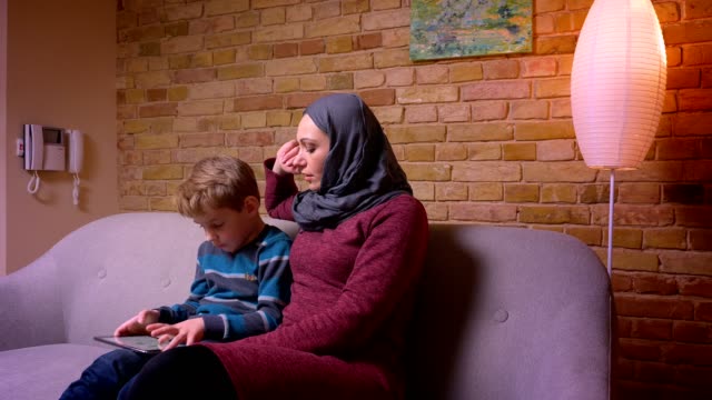 Konzentrierter-kleiner-Junge,-der-auf-dem-Tablet-spielt,-und-seine-muslimische-Mutter-im-Hijab,-die-seine-Aktivitäten-zu-Hause-beobachtet.