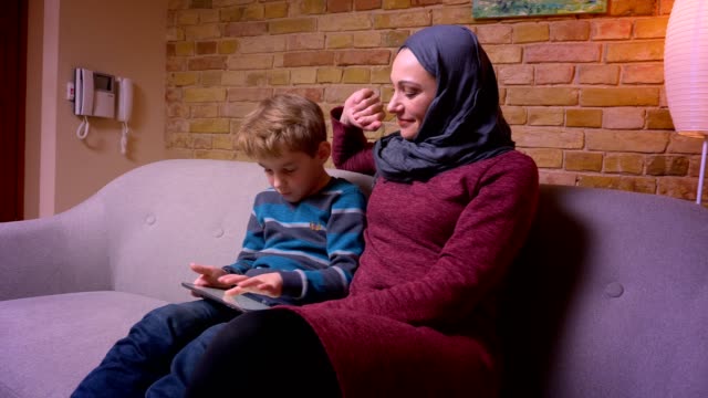 Konzentrierter-kleiner-Junge-spielt-Spiel-auf-Tablet-und-seine-muslimische-Mutter-in-Hijab-streichelt-ihn-zärtlich-zu-Hause.