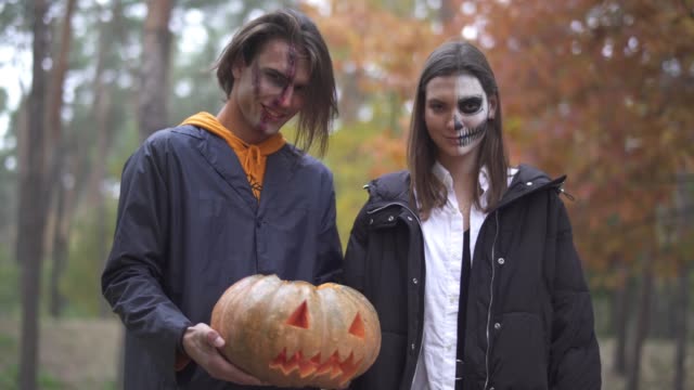 Tall-handsome-man-holding-Halloween-pumpkin-with-a-light-inside,-pretty-girl-standing-near,-both-smiling-then-making-evil-faces.