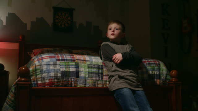 Sad-young-boy-stands-at-the-foot-of-his-bed-with-crossed-arms