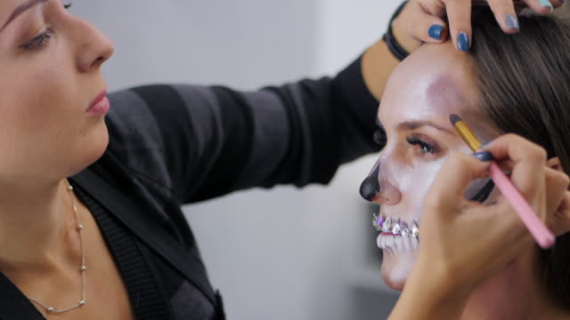 Makeup-artist-paints-greasepaint-for-Halloween-in-studio.-Woman-drawing-a-glamorous-skull-with-rhinestones-and-sequins-on-a-beautiful-young-girl-with-long-hair.-Slow-motion