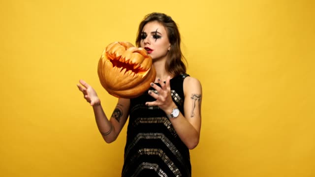 Young-nervous-displeased-lady-with-scary-make-up-playing-with-pumpkin-and-waiting-isolated-over-yellow