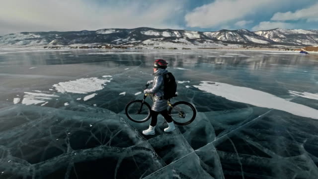 Woman-is-walking-beside-bicycle-on-the-ice.-The-girl-is-dressed-in-a-silvery-down-jacket,-backpack-and-helmet.-Ice-of-the-frozen-Lake-Baikal.-The-tires-on-the-bicycle-are-covered-with-special-spikes.