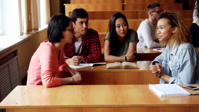 Studierende-diskutieren-Teamprojekt-sitzen-am-Schreibtisch-in-der-Universität,-Mädchen-und-jungen-sprechen,-Notizbücher,-Stifte-und-Smartphones-sind-sichtbar.-Teamwork-und-Kommunikation-Konzept.