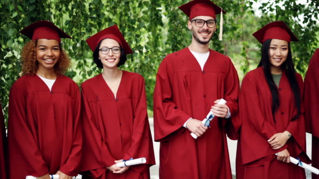 Dolly-shot-of-smiling-graduates-standing-in-line-on-campus,-looking-at-camera-and-holding-diplomas.-Bright-gowns-and-mortar-boards,-trees-and-scrolls-are-visible.