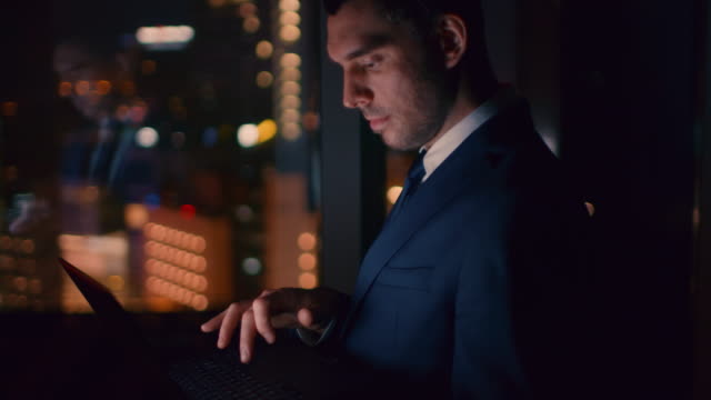 Late-at-Night-in-the-Office-Successful-Businessman-Holds-Laptop-while-Working-on-it.-In-the-Window-Business-District-View-with-City-Lights.