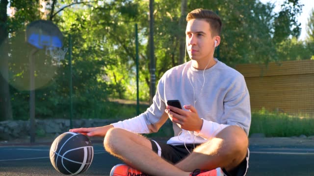 Junger-Mann-sitzt-auf-dem-Basketballplatz-und-Ball-dribbeln,-hören-Musik-über-Kopfhörer-und-Telefon-halten,-öffnen-Sie-Luft-und-Lens-flare