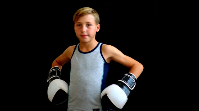 A-young-athlete-stands-on-a-dark-background.-The-boy-wears-a-gray-T-shirt-and-his-hands-in-gloves.-Black-and-white-gloves-are-visible-partially.-The-boy-has-blond-hair-and-dark-eyes