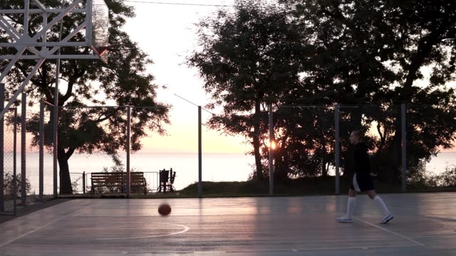 Stedicam-footage-from-the-side-of-a-young-girl-make-a-shot-to-the-basketball-net.-Outdoors,-trees-and-sun-shines-on-the-background