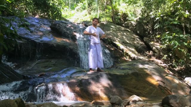Pensive-man-in-traditional-white-clothes-practices-chi-kung-exercises-standing-on-rock-near-falls.